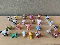 Photo showing a collection of 23 small Littlest Pet Shop animal figurines arrayed on a wood surface against a gray wall. Animals include pandas, foxes, owls, raccoons, dogs, birds, lizards, elephants, and hedgehogs in various pastel and brown colors.