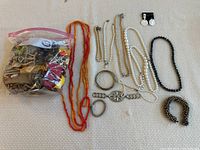 Jewelry items laid on white surface including orange-red beaded necklace, white pearl-like necklace, black beaded bracelet, silver-tone rings, earrings, and brooch, and a clear plastic bag filled with assorted jewelry bits.