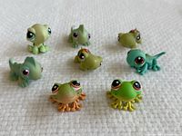Top-down view of all eight Littlest Pet Shop figures including frogs and dinosaurs on white fabric background.