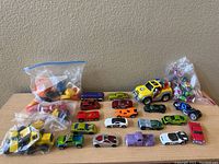 Photo showing collection of toy cars of different models and colors arranged on table along with bags containing toy figures and parts.