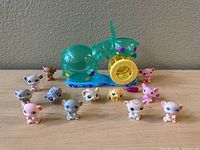 Photo of 10 small Littlest Pet Shop hamster figures arranged around teal hamster wheel accessory on beige surface against gray wall.