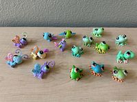 Photo showing 11 Littlest Pet Shop frog figures in varying shades of green and 5 winged insect-like figures with colorful translucent wings arranged on a flat surface.