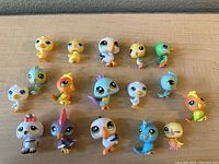 Top-down photo showing 16 Littlest Pet Shop toy bird figurines in various colors and styles, arranged on a wooden surface.