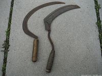 Pair of antique sickles laid on concrete surface showing overall shape and condition including curved blades and wooden handles.