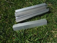Group of stacked aluminum architectural angle pieces on grass, showing multiple pieces with a flat metal finish and uniform shape.
