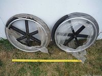 Two large black plastic wheels with six spokes each leaning against a white wall on a grassy surface. Both wheels have torn plastic wrapping and some tape residue. A yellow measuring tape at the base shows approximately 26 inches in diameter.