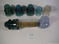 Photo of seven antique glass electrical insulators arranged in a row including one mounted on wood, showing a variety of green, aqua blue, clear, and light blue colors. The small light blue one has a damaged piece.