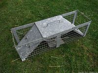 Side-angle view of the galvanized metal live trap on grass, showing wire mesh sides, solid metal top with handle, and angled metal panels at ends.