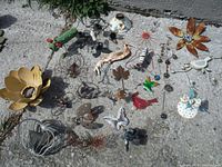 Various metal garden decor pieces arranged outdoors on stone, showing multiple flower shapes, animals, and whimsical figures with wear and rust.