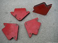 Four red welding magnets with angled shapes and screws visible, showing signs of light wear and used placed on concrete.