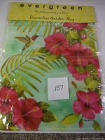 Photo of the garden flag folded in original packaging showing pink hibiscus flowers, green leaves and a hummingbird with brand name Evergreen on yellow header.