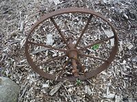 Top-down view of the rusty antique metal wheel showing spider-like metal spokes and the central axle lying on ground covered with wood chips.