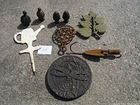 Overview photo showing all nine cast iron pieces arranged on concrete surface: three finials, watering can shape, leaf trivet, small shovel, round dragonfly trivet, and decorative plate.