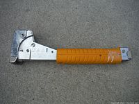 Side profile of metal hammer stapler with orange grip handle on concrete floor.