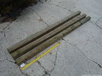 Four used 4x4 pressure treated wooden posts lying parallel on cracked concrete, a measuring tape beside them showing length over 7 feet.