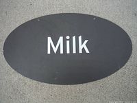 Front side of the oval rigid plastic sign with 'Milk' printed in white on black background, showing mounting holes.