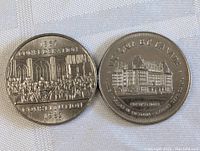 Two Canadian dollar coins side by side on a white cloth background. Left coin commemorates the 1867 Confederation Constitution 1932 with a historical scene. Right coin shows the Empress Hotel in Victoria, BC with detailed building engraving.