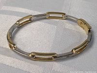 View of the sterling silver and gold tone linkage bracelet resting on fabric, showing the alternating open rectangular gold tone and solid silver links in a circular formation.