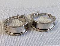 Pair of hoop earrings shown lying flat on light fabric, highlighting the detailed surface texture and rope edges.