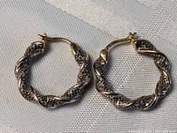 Pair of sterling silver twisted hoop earrings with textured encrusted detailing placed on fabric surface, showing overall design and latch back closure.