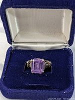 Front angled view of rectangular faceted purple gemstone set on a sterling silver ring with rope detail on band, shown in blue velvet jewelry box with cream satin top.