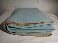 Folded light blue wool blanket showing orange edge stitching on a plain white surface and wooden panel background.