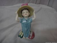 Front view of Royal Doulton figurine of a girl in blue dress holding a large hat with pink flowers, with shoes and handbag at her feet.