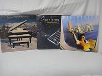 Front covers of three Supertramp vinyl albums: Even in the Quietest Moments (broken piano in snowy landscape), Crime of the Century (window bars and stars), Breakfast in America (waitress with glass and city skyline).