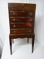 Front view of a dark wooden jewelry cabinet with brass handles on five drawers and standing on four legs.
