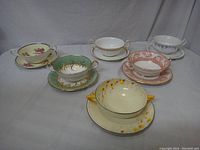 Photo showing eight double handled tea cups and various matching saucers arranged on fabric background. Assorted floral and decorative patterns. One saucer appears damaged.