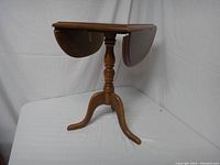 Wooden drop leaf side table shown with leaves down revealing a smaller table surface and central pedestal base with three curved legs.