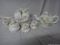 Full view of the ceramic tea set showing teapot, sugar bowl, creamer, cups, and saucers all with embossed pink rose and leaf details.