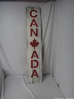 Front view of the wooden sign painted with 'CANADA' vertically and a red maple leaf symbol, standing against a white fabric background.