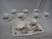 Seven floral bone china tea cups arranged behind a tray holding sugar bowl with spoon and creamer, all with a matching Lavender Rose floral pattern and gold trim.