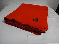Full folded view of the bright orange wool blanket with visible black edge stitching on a white surface.