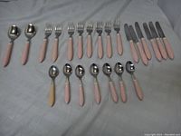 Entire cutlery set laid out, including soup spoons, forks, knives, and teaspoons with pink handles and one cream handled teaspoon visible.