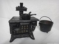Front and side angle view of the miniature metal wood stove with pot and ladle on the top and a cast iron pot beside it.