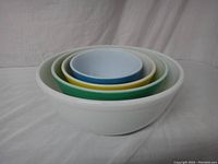 Four nested PYREX mixing bowls in blue, yellow, green, white aligned inside each other on white fabric.