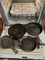 Four cast iron skillets arranged on open oven door with aluminum grid tray on shelf above