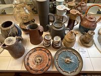 Full overview of pottery collection including wall clocks, pitchers, teapots, cups, and jars displayed on counter