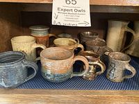 Front view of 9 handmade stoneware mugs showing various glazes and textures on a blue mat inside a wooden shelf.
