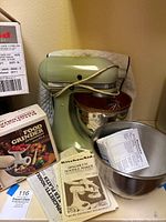 Green KitchenAid stand mixer with copper mixing bowl and stainless steel mixing bowl on side, along with boxed Food Grinder attachment and Spaghetti/Noodle Maker instruction booklet visible.