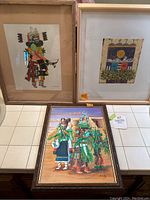 Photo showing three Native American artworks: two framed prints and one framed painting placed on and against a table surface
