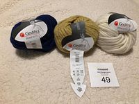Three balls of Gedifra Benevito yarn in blue, mustard yellow, and cream colors displayed on a beige textured surface.