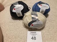 Three balls of Gedifra Extra Soft Merino Fino yarn in black, blue, and white colors lying on a textured cream surface.
