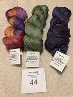 Three skeins of hand dyed Silky Sock yarn in Hummingbird, Rosemary, and Twilight Dance colors spread side by side on a beige background with product tags and auction card.