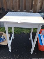 Small vintage table painted white with turned legs and plank top, photographed outdoors with grass and fence in background.