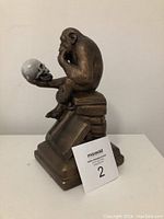 Front and side view of the bronze-colored ape statue sitting on books holding a skull with some chips visible.