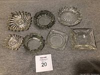 Photo showing 7 glass/crystal ashtrays of various shapes and textures placed on a grey surface. Ashtrays appear in very good condition with no chips.