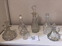 Photo showing 6 varying clear glass and crystal decanters with tops arranged on a white shelf.